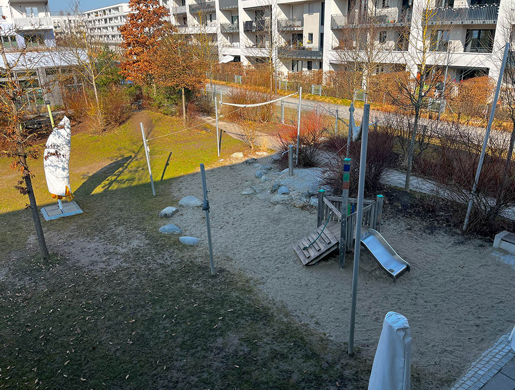 Spielplatz mit Rutsche, Klettergerüst und Sandfläche; umgeben von Wohngebäuden und Bäumen.