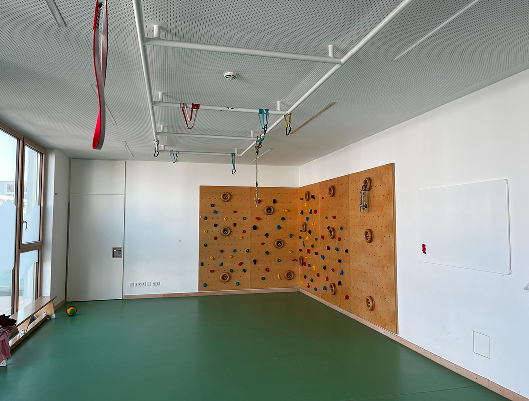 Kletterwand in heller Halle, bunter Griffmuster, grüne Matte, Gymnastikbänder an der Decke.