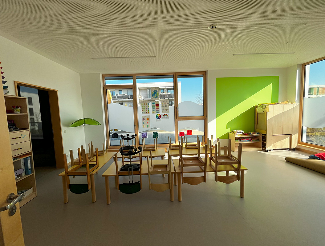 Ein leerer, heller Raum mit aufgestellten Stühlen, Spielzeugregal und grüner Wand.