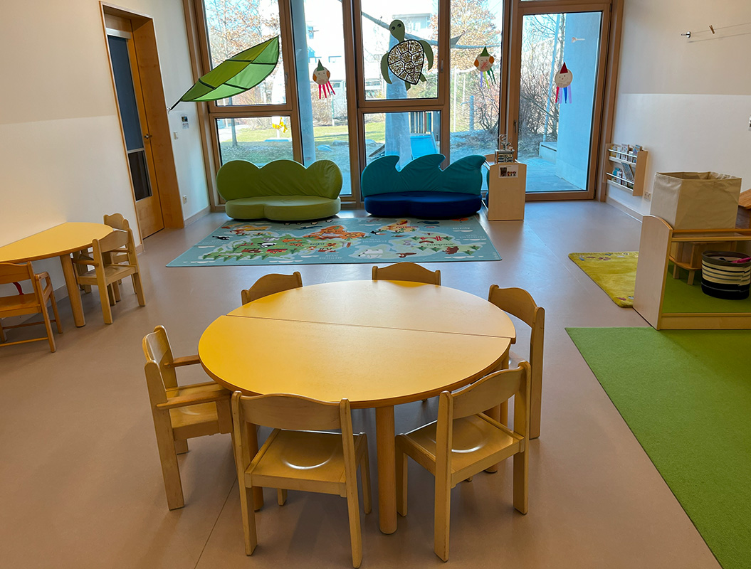 Ein heller Kindergartenraum mit runden Holztischen und bunten Blattform-Sofas.