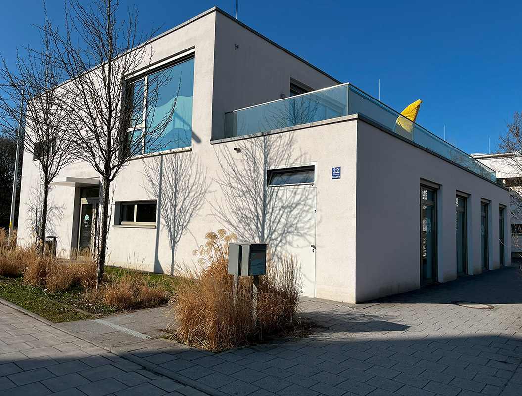 Modernes, weißes Gebäude mit Flachdach und Glasbalkon, umgeben von kahlen Bäumen und Gras.