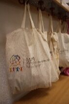 Mehrere beigefarbene Taschen hängen an Haken, eine ist mit buntem Logo verziert.