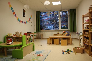 Ein bunter Kindergartenraum mit Tischen, Stühlen und Spielzeug auf hellem Boden.